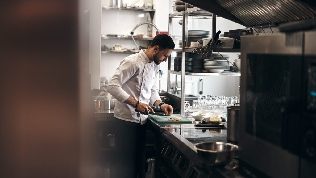A chef in a white coat chopping vegetables on a green board in a kitchen
